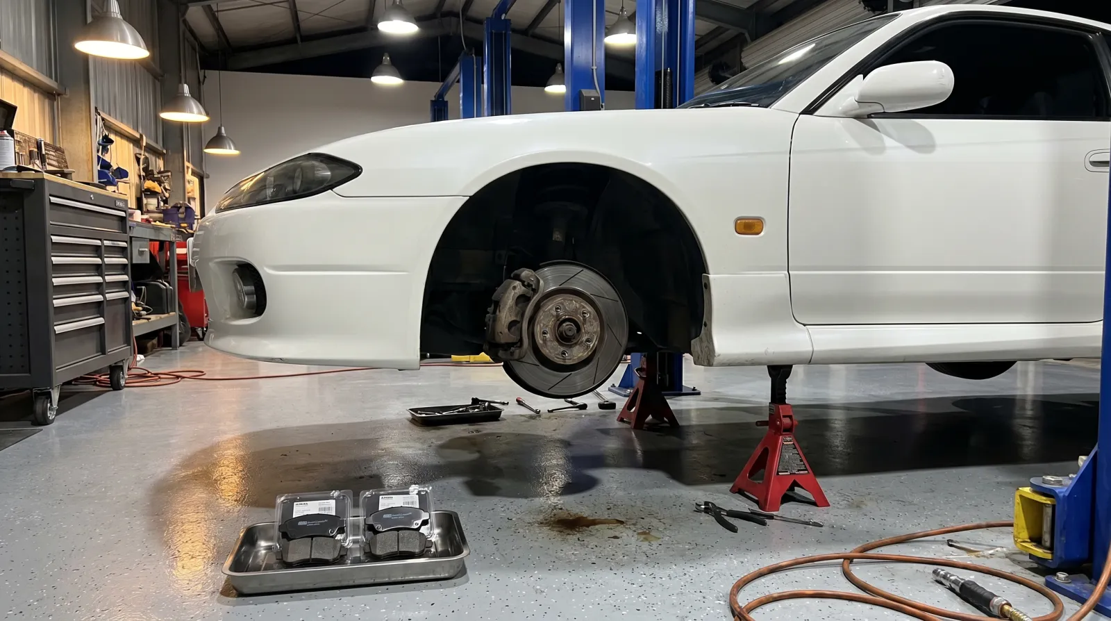 A disc brake caliper and rotor exposed on a lifted JDM coupe with new brake pads ready on the bench