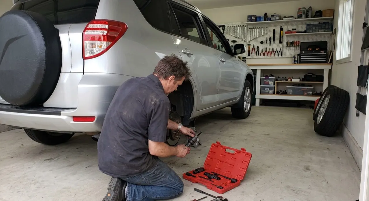 A mechanic using a brake caliper wind-back tool on a rear disc brake caliper, silver aluminum tool engaged in the piston slots, brake rotor and knuckle visible in background, workshop lighting, close-up detail
