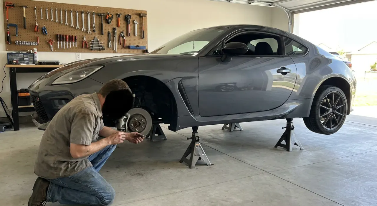 A floor jack positioned beneath a car chassis in a clean concrete garage, with two red jack stands placed at the correct subframe points and the car visibly resting on the stands, floor jack slightly lowered
