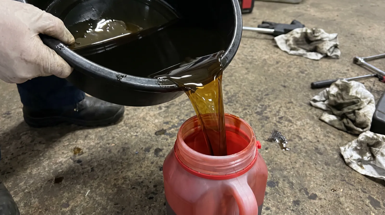 Used engine oil being poured from a drain pan into a sealable jug on a garage floor