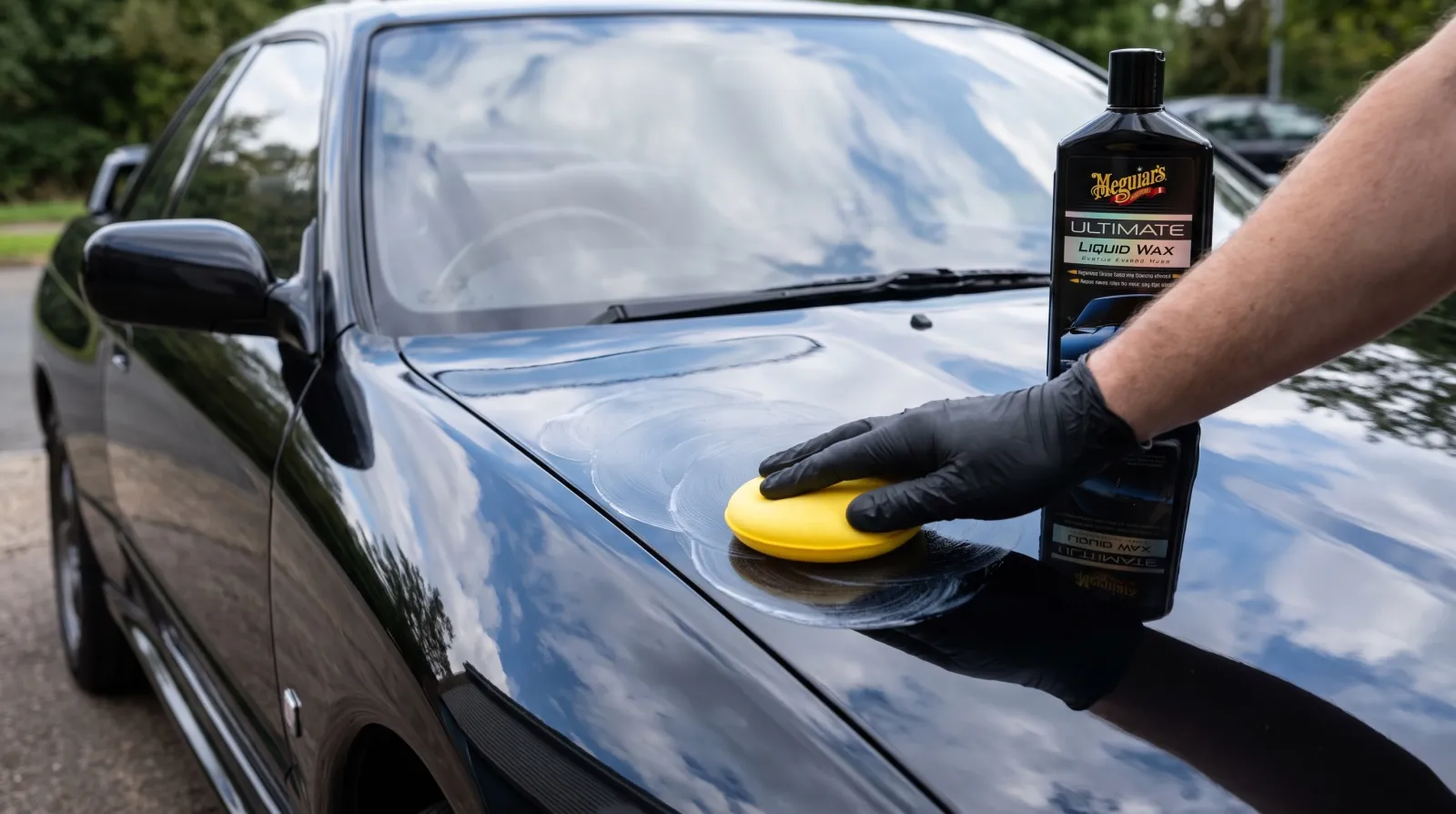 Liquid car wax being worked into the glossy dark hood of a sports coupe with a yellow foam applicator